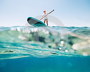 Under the water view angle to the smiling blonde teenager, boy rowing stand up paddleboard. Active family summer vacation time