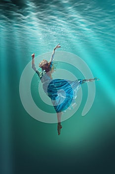 An underwater ballet. Beautiful young girl in elegant dress gracefully levitating underwater with sunlight above.