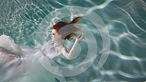 Underwater girl. Beautiful red-haired woman in a white dress, swimming under water.