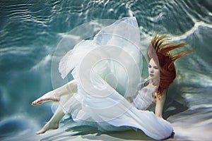 Underwater girl. Beautiful red-haired woman in a white dress, swimming under water.