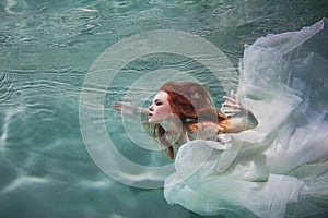 Underwater girl. Beautiful red-haired woman in a white dress, swimming under water.