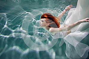 Underwater girl. Beautiful red-haired woman in a white dress, swimming under water.