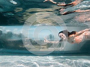 Underwater photo of aisan woman in bikini diving in the swimming pool with transparent glass wall. infinity pool edge on tropical