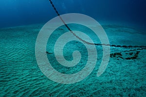 Underwater photo of heavy steel anchor chain that hangs down from water surface and lays on white sand bottom, murky blue water