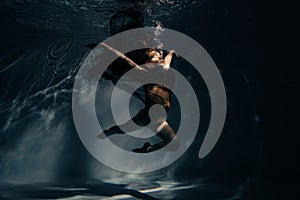Underwater shoot of ballerina in black dress swimming and dancing in water through sunbeams