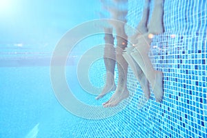 Underwater Shot Of Couples Legs As They Sit On Edge Of Swimming Pool On Vacation