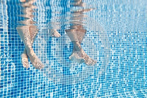 Underwater Shot Of Couples Legs As They Sit On Edge Of Swimming Pool On Vacation