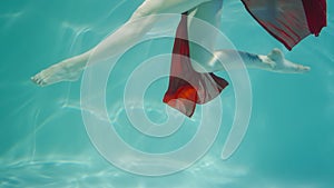 Underwater shot of legs woman dancing in red chiffon costume. Female dancer moves in blue water column with glare