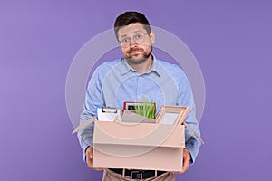 Unemployed man with box of personal office belongings on purple background