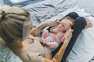 Unexperienced young mother with tattoos on her forearms sitting calmly on bed, holding her small infant baby boy on her