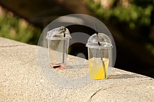 Unfinished chilled drinks in plastic disposable glasses are thrown on a stone fence in the park. Orange and strawberry juice with