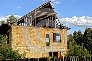 Unfinished house of brown brick and wooden roof among the green trees behind the fence