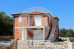 Unfinished red bricks family house with new doors and windows surrounded with stone wall and building material with trees and