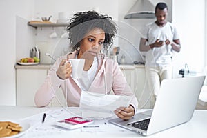 Unhappy african american woman drinking coffee, her husband calls bank to manage debt