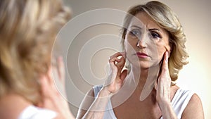 Unhappy aged woman looking in mirror at home, touching face, aging process