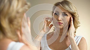 Unhappy aged woman looking in mirror at home, touching her face, aging process
