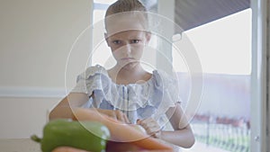 Unhappy angry little girl in front of plate with vegetables in the kitchen. The child pushes away a plate of tasteless