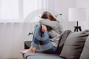 Unhappy anxiety young brunette woman 25s covering face with knee sitting on couch in living room at home, hurt heartbroken female