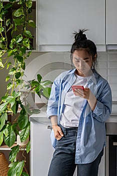 Unhappy Asian girl standing in kitchen at home holding cellphone spying on boyfriend in social media