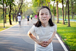 Unhappy asian little cute girl holding her hands on belly,suffers from stomach pain,colic after running,exercise in outdoor park, photo