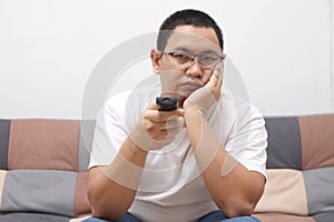 Unhappy Asian man at home bored and frustrated watching tv movie on sofa couch holding remote control