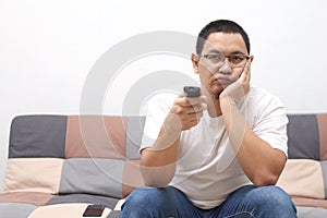 Unhappy Asian man at home bored and frustrated watching tv movie on sofa couch holding remote control