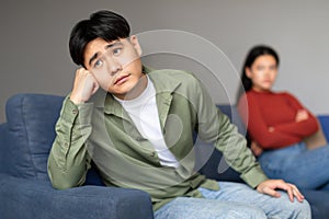 Unhappy Asian Man Sitting Separate From Discontented Wife At Home