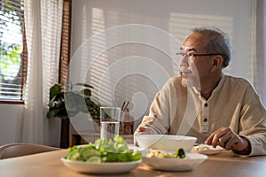 Unhappy Asian Senior older man sit alone, eat foods on table in house. Depressed mature attractive elderly retired grandfather