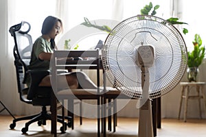Unhappy Asian woman sitting in front of fan at work suffering from heat in modern house