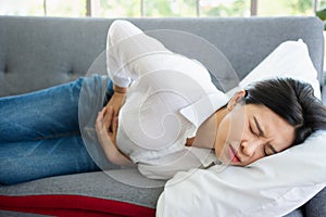 Unhappy Asian woman sitting on the sofa and holding on stomach suffering. Abdominal pain that comes from menstruation, diarrhea,