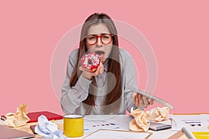 Unhappy attractive young woman cries as has problems at work, eats tasty sweet donut, carries modern touchpad, develops