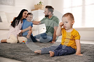 Unhappy baby sitting alone on floor while parents spending time with his elder brother. Jealousy in family