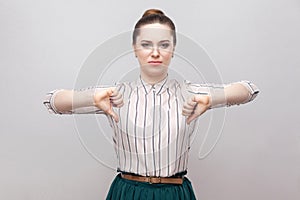 Unhappy beautiful young woman in striped shirt and green skirt with makeup and collected ban hairstyle, standing with thumbs down