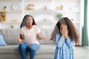 Unhappy black girl covering face with hands, crying while mom scolding her at home, empty space