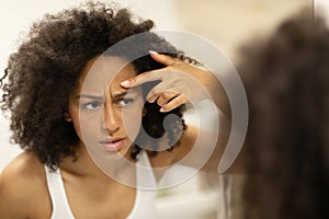 Unhappy black woman with problem skin squeezing a pimple on her forehead, suffering from acne
