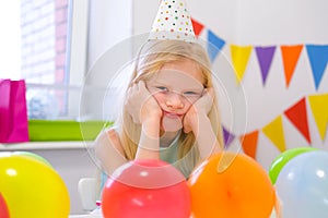 Unhappy blonde caucasian girl with boring face near birthday rainbow cake. Festive colorful background. Bad birthday