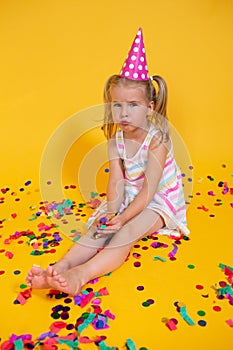 Unhappy blonde caucasian girl with boring face with scattered confetti. Yellow studio background. Bad birthday party