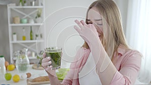 Unhappy Caucasian girl closing nose with fingers to drink green healthful cocktail. Portrait of slender young blond