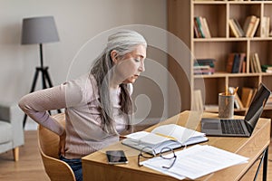 Unhappy caucasian old female with gray hair works on computer and suffers from back pain in room interior