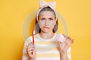 Unhappy confused woman holding marshmallow and toothbrush, dressed in casual striped t shirt isolated over yellow background,