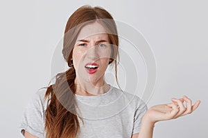 Unhappy confused young woman with red hair and freckles looks displeased and holding copyspace on palm isolated over white