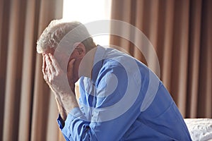 Unhappy And Depressed Senior Man With Head In Hands Sitting On Edge Of Bed At Home