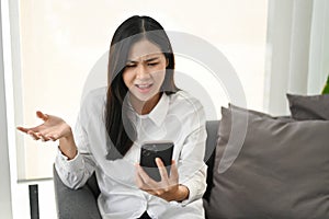 Unhappy and dissatisfied Asian woman sitting on sofa, receiving unreasonable news or message