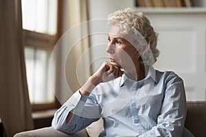 Unhappy elderly female sitting on couch propping chin with hand