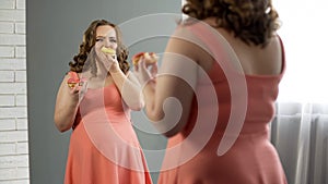 Unhappy fat female eating donuts in front of mirror, nutrition disorder issue