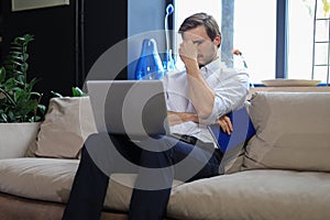 Unhappy frustrated young male holding head by hand sitting with laptop on sofa.