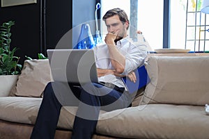 Unhappy frustrated young male holding head by hand sitting with laptop on sofa.