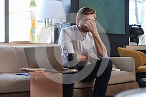 Unhappy frustrated young male holding head by hand sitting with laptop on sofa.