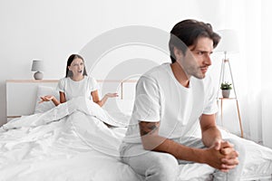 Unhappy frustration european young man ignoring angry woman, lady sits in bed and scream on guy in white bedroom
