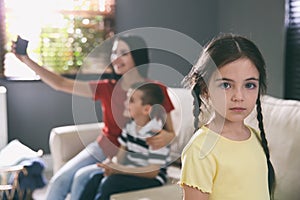 Unhappy girl feeling jealous while mother taking selfie with her brother at home